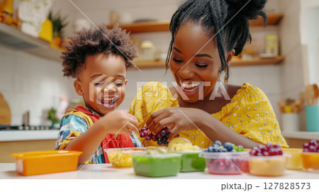 Black mom and preschool son joyfully preparing lunch boxes in bright kitchen with healthy food. Clean and bright kitchen background 127828573
