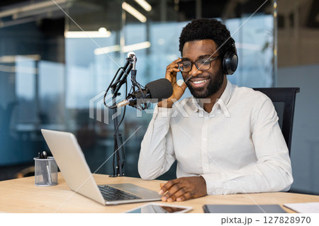 Smiling African American man wearing headphones, sitting at a desk with a microphone and laptop during a podcast recording. Smiling African American man wearing headphones, sitting at a desk with a microphone and laptop during a podcast recording. 127828970