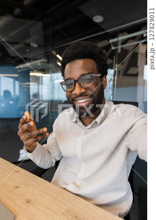 A smiling man in glasses is gesturing, likely in a video call, showcasing enthusiasm in a modern office setting A smiling man in glasses is gesturing, likely in a video call, showcasing enthusiasm in a modern office setting 127829011