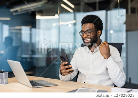 Successful businessman celebrating victory and triumphant achievement results. Man with phone in hands sitting at desk with laptop inside office 127829025