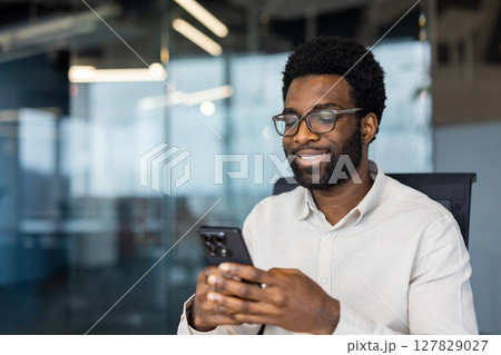 A smiling man wearing glasses looks at his phone while sitting in a modern office. It seems he is reading or watching something interesting. 127829027