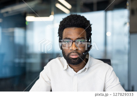 Portrait of a young man with a headset, a support worker looking thoughtfully and seriously at the camera, a member of a customer service team, an online consultant inside an office. Portrait of a young man with a headset, a support worker looking thoughtfully and seriously at the camera, a member of a customer service team, an online consultant inside an office. 127829333