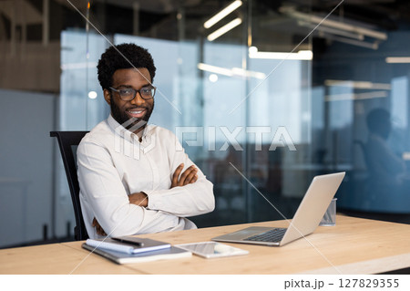Portrait of a young happy entrepreneur, businessman smiling and looking at the camera, sitting at a desk with a laptop inside a company office, man in business clothes. 127829355