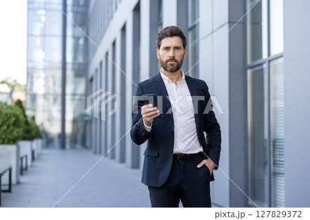 A well-dressed businessman stands confidently, using a smartphone against an office building backdrop, ready for communication. A well-dressed businessman stands confidently, using a smartphone against an office building backdrop, ready for communication. 127829372