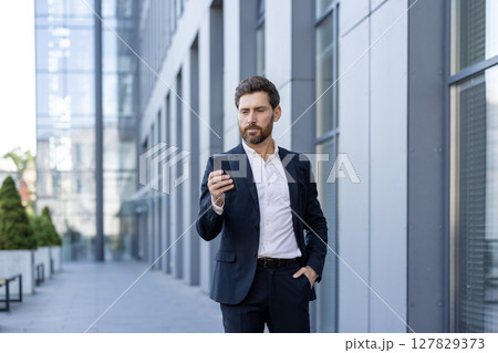 A man in a suit stands outside a building, absorbed in his phone. He holds his hand in his pocket, focused on the screen as he checks his messages. A man in a suit stands outside a building, absorbed in his phone. He holds his hand in his pocket, focused on the screen as he checks his messages. 127829373