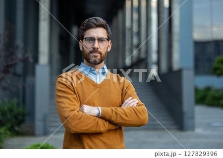 A confident man with glasses stands with arms crossed in front of a modern building, outdoors. A confident man with glasses stands with arms crossed in front of a modern building, outdoors. 127829396