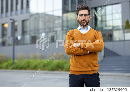 Confident businessman poses in front of a modern building. He wears glasses and an orange sweater, exuding professional confidence and success. Confident businessman poses in front of a modern building. He wears glasses and an orange sweater, exuding professional confidence and success. 127829406