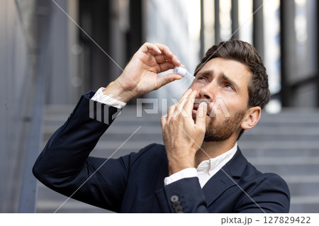 A businessman applies eye drops to soothe irritated eyes. He stands outdoors near office buildings, showing the challenges of modern life. A businessman applies eye drops to soothe irritated eyes. He stands outdoors near office buildings, showing the challenges of modern life. 127829422
