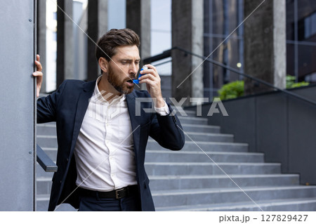 A professional man uses an inhaler outside of a modern office building. He appears to be struggling with a sudden asthma attack or breathing problem while on the stairs. A professional man uses an inhaler outside of a modern office building. He appears to be struggling with a sudden asthma attack or breathing problem while on the stairs. 127829427