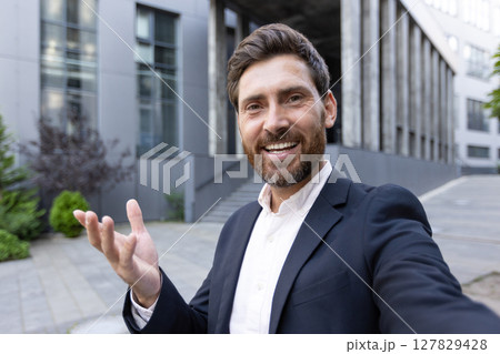 A businessman in a video call, smiling and gesturing outdoors in front of a building A businessman in a video call, smiling and gesturing outdoors in front of a building 127829428