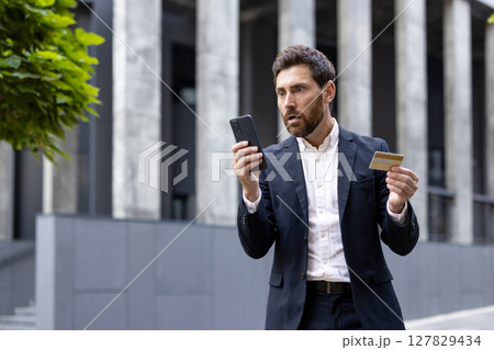 A businessman looks shocked while holding a credit card and his phone, possibly due to a financial issue. 127829434