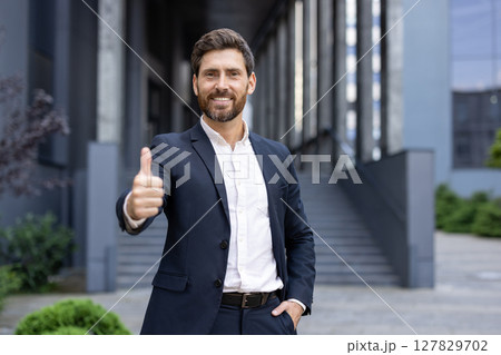 A smiling businessman in a suit, giving a thumbs-up gesture while standing outside of a modern building. 127829702