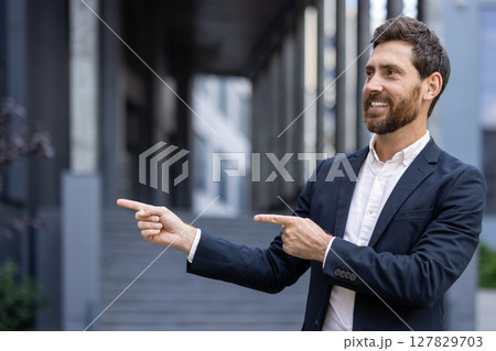 A smiling businessman in a dark suit points both fingers to the right, near an office building, ready to lead. 127829703