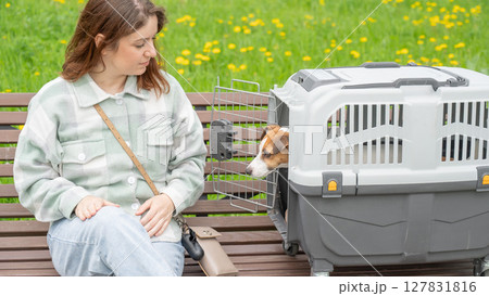 Caucasian woman traveling with dog. Owner sits on bench with Jack Russell Terrier in cage for safe travel. Caucasian woman traveling with dog. Owner sits on bench with Jack Russell Terrier in cage for safe travel. 127831816