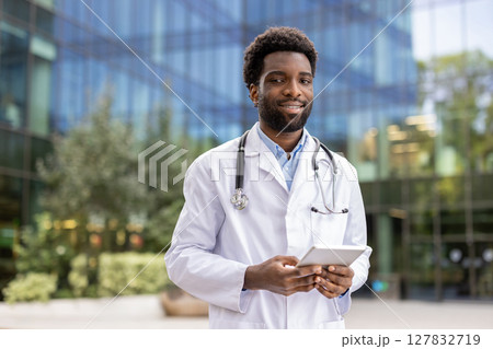 A smiling doctor in a white coat holds a tablet computer, appearing confident and professional in an outdoor setting with a building in the background. A smiling doctor in a white coat holds a tablet computer, appearing confident and professional in an outdoor setting with a building in the background. 127832719