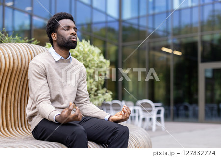 An african american man meditates peacefully outdoors, finding calm in the natural light near a modern building. 127832724