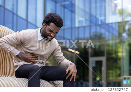 Man suffering from stomach pain, businessman sitting on a bench outside an office building holding hands by his side, patient has inflammation and poor digestion. Man suffering from stomach pain, businessman sitting on a bench outside an office building holding hands by his side, patient has inflammation and poor digestion. 127832747