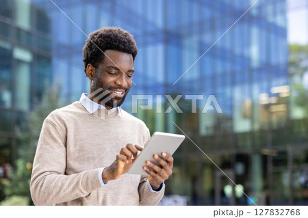 A smiling businessman looks at his tablet while standing outdoors near a modern office building. A smiling businessman looks at his tablet while standing outdoors near a modern office building. 127832768