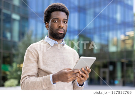 An african american man in a light sweater, holding a tablet, posing in front of an office building, looking at the camera. An african american man in a light sweater, holding a tablet, posing in front of an office building, looking at the camera. 127832769