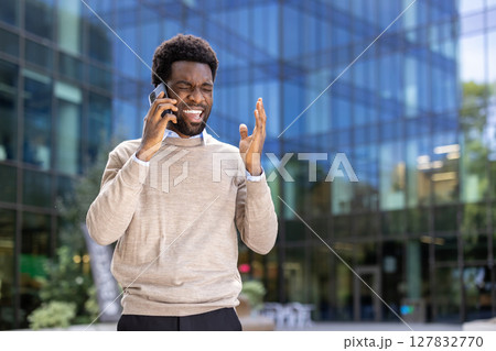 An african-american businessman looks stressed while having a phone call outdoors, in front of a modern building. He is visibly angry. An african-american businessman looks stressed while having a phone call outdoors, in front of a modern building. He is visibly angry. 127832770