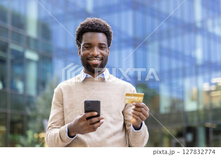 A cheerful man holding a smartphone and credit card, smiling towards the camera in an outdoor setting. He is likely engaged in a transaction. 127832774