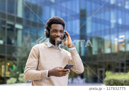 Smiling man wearing headphones holds a phone in front of a modern building with a glass facade. Smiling man wearing headphones holds a phone in front of a modern building with a glass facade. 127832775