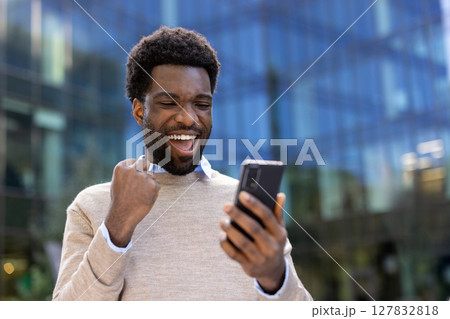 A joyful black man reacts with excitement while holding a smartphone, likely celebrating a win outdoors. 127832818