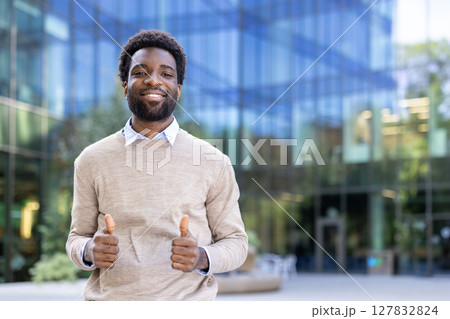 A smiling black man gives a thumbs up, standing in front of a modern building. The scene conveys approval and positivity. 127832824