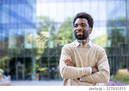 A smiling, confident businessman stands outside with his arms crossed, exuding success in a modern cityscape. 127832835