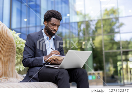 Focused businessman in a suit works outdoors on a laptop while jotting notes, wearing earbuds against a modern building backdrop. Focused businessman in a suit works outdoors on a laptop while jotting notes, wearing earbuds against a modern building backdrop. 127832877
