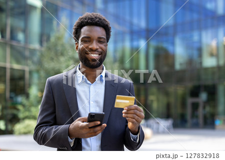 Joyful businessman with bank credit card and phone outside office building. Man smiling and looking at camera, satisfied with online shopping and money transfer remotely. Joyful businessman with bank credit card and phone outside office building. Man smiling and looking at camera, satisfied with online shopping and money transfer remotely. 127832918