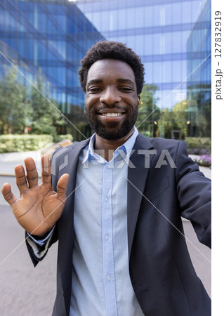 A young, handsome man in a blazer smiles and waves to the camera during a mobile video call in front of a modern office building 127832919