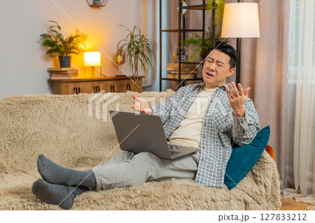 Upset displeased Chinese man using laptop computer reading bad news sitting on sofa in living room Upset displeased Chinese man using laptop computer reading bad news sitting on sofa in living room 127833212