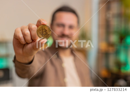 Portrait of happy young male freelancer trader showing golden bitcoins sitting at home office table 127833214