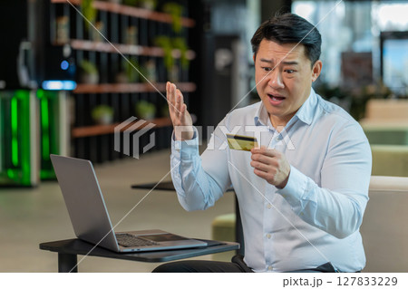 Angry Chinese mature businessman holding credit bank card sitting at modern workplace in office Angry Chinese mature businessman holding credit bank card sitting at modern workplace in office 127833229