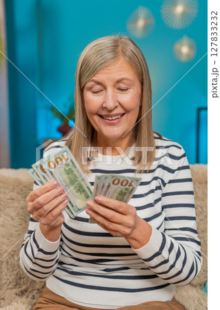 Mature woman joyfully counting dollar money on sofa after lottery win, happy with bonus gift at home 127833232