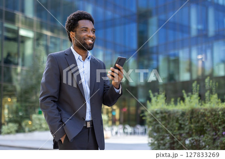 A smiling african american businessman looks at his phone outside a modern office building on a sunny day. A smiling african american businessman looks at his phone outside a modern office building on a sunny day. 127833269