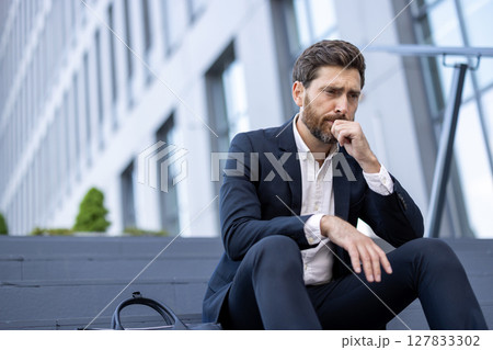 A pensive businessman in a suit sits outside, deep in thought. His expression suggests stress or worry in a corporate environment. A briefcase sits nearby. A pensive businessman in a suit sits outside, deep in thought. His expression suggests stress or worry in a corporate environment. A briefcase sits nearby. 127833302