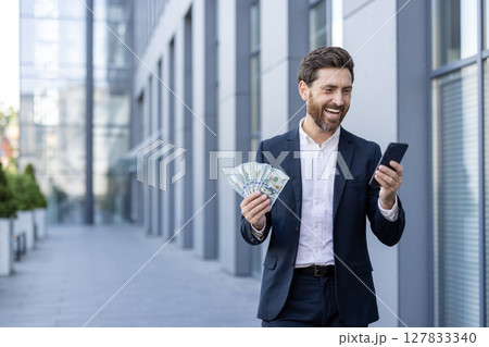 A smiling businessman in a suit holds cash and his phone, possibly celebrating a financial success outside the office. A smiling businessman in a suit holds cash and his phone, possibly celebrating a financial success outside the office. 127833340