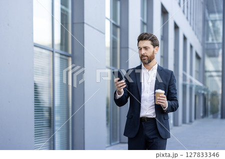 A man in a suit reviews his phone while walking outdoors, holding a coffee cup. The background shows a modern office building. 127833346