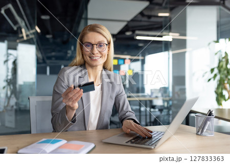 A blonde businesswoman in a gray suit smiles while holding a credit card and using her laptop in a modern office setting. A blonde businesswoman in a gray suit smiles while holding a credit card and using her laptop in a modern office setting. 127833363