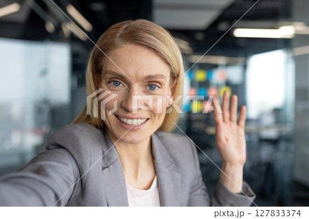 A smiling blonde businesswoman waves hello during a video call, with a professional office setting in the background. 127833374