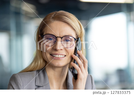 A smiling businesswoman with glasses holds a phone near her ear, looking directly at the camera in a professional setting. 127833375
