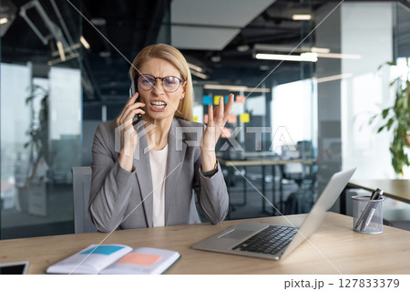 An angry businesswoman in a modern office is on a phone call, looking frustrated and stressed. She is gesturing with her hand and wearing glasses. An angry businesswoman in a modern office is on a phone call, looking frustrated and stressed. She is gesturing with her hand and wearing glasses. 127833379