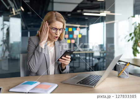 A shocked businesswoman reacts to information on her phone while working at her desk in the office. 127833385