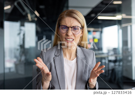 Video call, upset and disappointed woman looking into webcam, talking to colleagues and partners dissatisfied sitting inside office, online business meeting conference. 127833416
