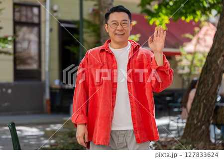 Asian man smiling friendly at camera, waving hands hello, hi, greeting or goodbye in city street 127833624