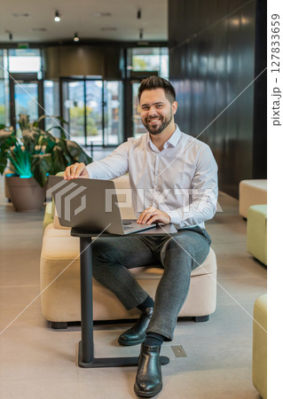 Young Caucasian businessman in while shirt opens laptop and starts working sitting in office lobby 127833659