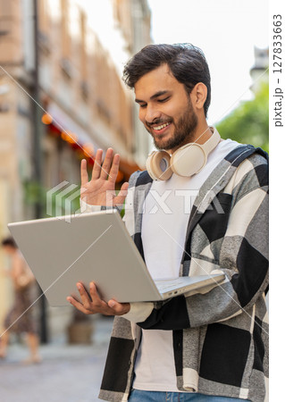 Young Indian man freelancer sitting on street, looking at laptop camera, making video call outdoors 127833663