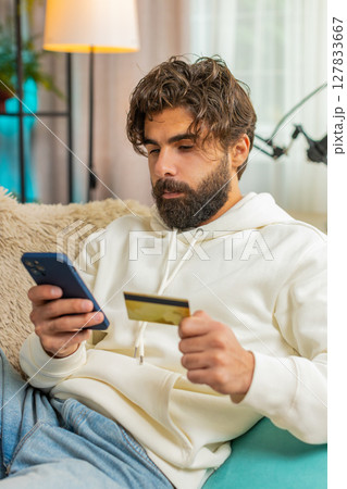 Happy young Hispanic man guy using credit bank card and cellphone while shopping online at home Happy young Hispanic man guy using credit bank card and cellphone while shopping online at home 127833667